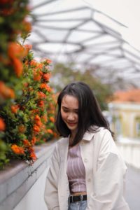 shy young asian woman wearing an open white blouse standing next to flower bushes